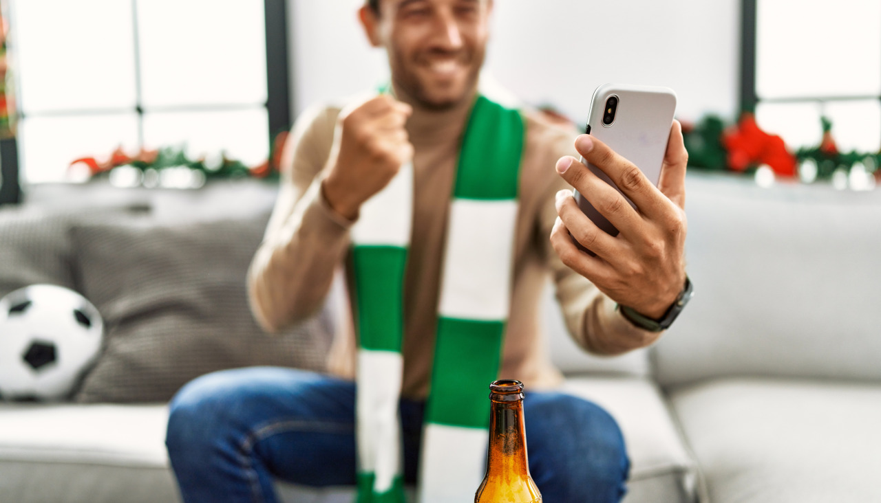 Young hispanic man supporting soccer match using smartphone sitting by christmas decor home
